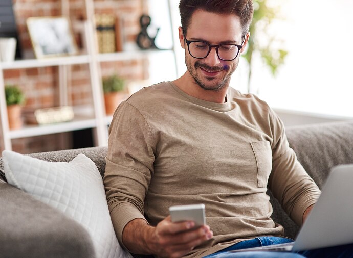 Mann sitzt auf Sofa mit Notebook auf dem Schoß und schaut auf sein Mobiltelefon